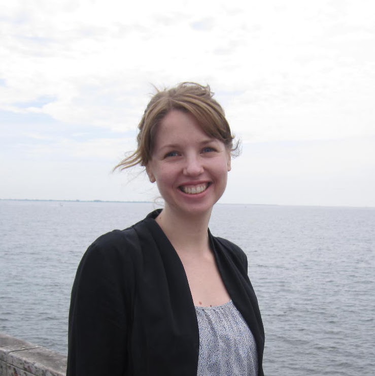 A woman smiling, posing in front of the sea in a cardigan and dotted top.