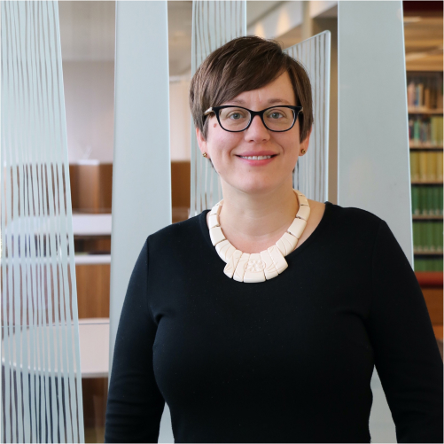 Light-skinned woman with brown hair wearing glasses and a chunky necklace in a library.